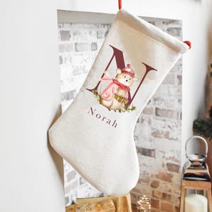 May include: A cream-colored Christmas stocking featuring a large maroon letter N, a watercolor illustration of a small animal wearing a pink hat and scarf, and the name Norah printed in maroon text below the graphic on the front of the stocking.