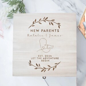 May include: A wooden box with a light brown stain. The box has an engraved design of a leafy wreath, a stylized baby, and the text "NEW PARENTS Natalie & James EST. 2024 LET THE ADVENTURE BEGIN".