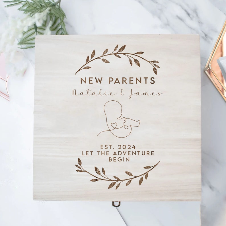 May include: A wooden box with a light brown stain. The box has an engraved design of a leafy wreath, a stylized baby, and the text "NEW PARENTS Natalie & James EST. 2024 LET THE ADVENTURE BEGIN".