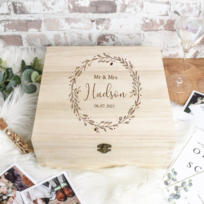 May include: A light-colored wooden box with a hinged lid, featuring a decorative wreath design and the engraved text "Mr & Mrs Hudson 06.07.2021". The box has a metal clasp and is likely for keepsakes or gifts.