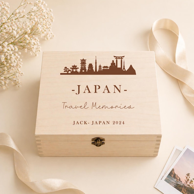 May include: A light-colored wooden box with a hinged lid. The lid features a brown silhouette of Japanese landmarks, the word "-JAPAN-", and the text "Travel Memories". The box is secured with a metal clasp and includes the text "JACK - JAPAN 2024".