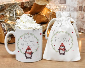 Może przedstawiać: Biały kubek ceramiczny z wzorem czerwonego i zielonego pingwina i tekstem "Millie's Christmas Hot Chocolate". Obok kubka znajduje się biała torba sznurkowa z wzorem czerwonego i zielonego pingwina i tekstem "Millie".