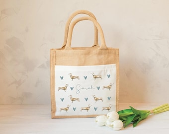 May include: A tan jute tote bag with two handles and a white front pocket. The pocket features a repeating pattern of brown dachshunds and blue hearts, with the name Sarah printed in blue script in the center of the design.
