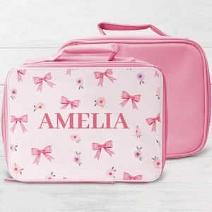 May include: A pink lunch bag and a smaller matching bag. The front of the lunch bag is decorated with pink bows, floral patterns, and the name "AMELIA". The smaller bag is a solid pink colour.