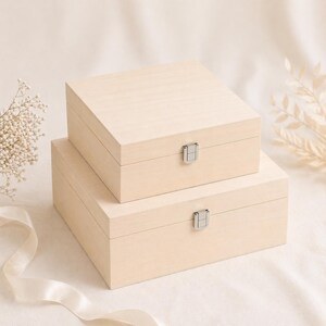 May include: Two unfinished wooden boxes, one stacked on top of the other. Each box has a rectangular shape with a hinged lid and a silver-colored metal clasp. The boxes are light-colored and appear to be made of natural wood.