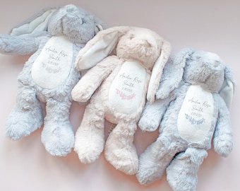 May include: Three plush rabbit toys with soft fur in shades of light gray, cream, and blue. Each rabbit features a white oval patch on its chest printed with the name Amelia Rose Smith, a date, and a small floral graphic in matching colors.