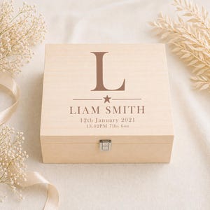 May include: A light-coloured wooden keepsake box with a hinged lid and a silver-coloured clasp. The lid features the letter "L", the name "LIAM SMITH", and birth details: 12th January 2021, 13.02PM, 32.2 kg.