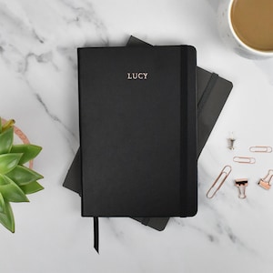 May include: A black notebook with the name "LUCY" in rose gold lettering. The notebook has a black elastic band and a black ribbon bookmark. A second black notebook is partially visible underneath. Other items include paperclips, a drawing pin, a succulent, and a cup of coffee.