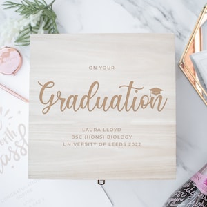 May include: A wooden box with a gold inscription that reads "On Your Graduation" and includes the name Laura Lloyd, her degree, and the year 2022. The box is surrounded by other graduation gifts, including a bottle of champagne, a small box, and a makeup brush set.