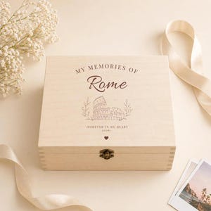 May include: A square light-colored wooden keepsake box with a metal latch. The lid features the text "MY MEMORIES OF Rome" above a line drawing of the Colosseum, the text "FOREVER IN MY HEART 2026", and a small heart icon.