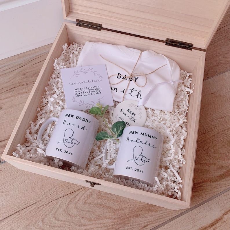 May include: A wooden gift box filled with baby shower gifts. The box contains two white mugs with the text "New Daddy" and "New Mummy" with names and the year 2024. There is also a white onesie with the text "Baby Smith" and a small round ornament with the text "Baby Smith May 2024".