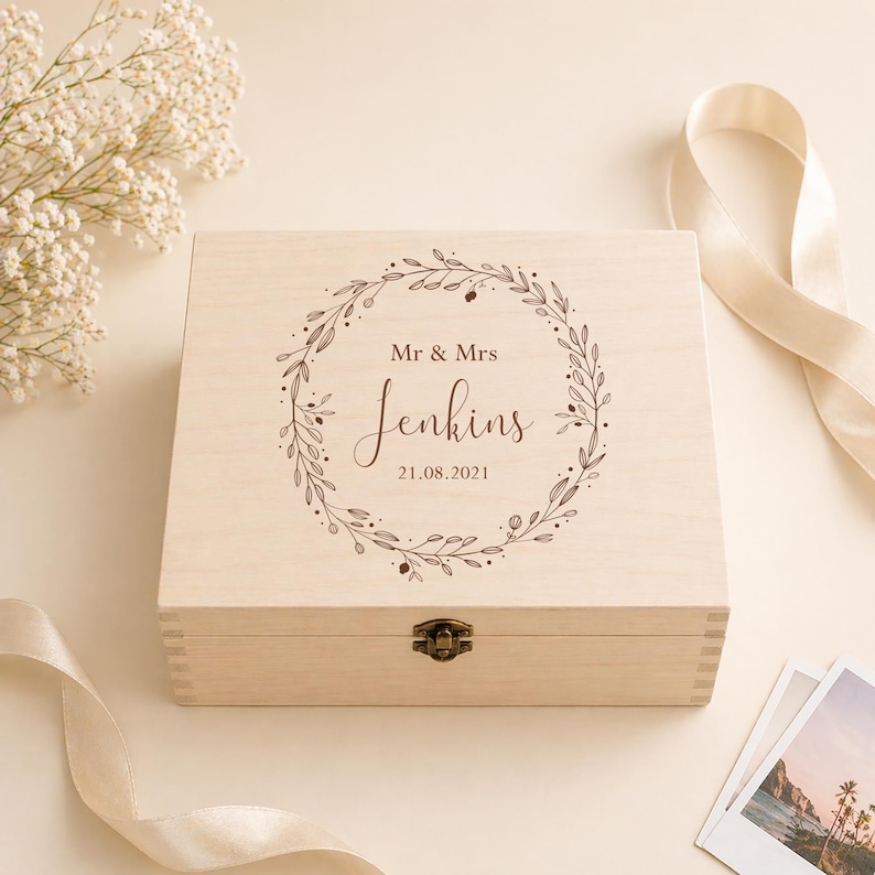 May include: A light-colored wooden box with a hinged lid and a metal clasp. The box is engraved with the words "Mr & Mrs Jenkins" and the date "21.08.2021" inside a decorative wreath. The box is surrounded by flowers and a ribbon.