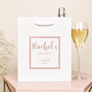 May include: A white gift bag with a pink square border and text that reads Rachel's HEN PARTY LONDON 2025. The bag features a white cord handle and is positioned next to a clear glass filled with a light yellow liquid and a bottle top.