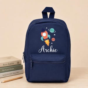 May include: A navy blue backpack featuring a graphic of an orange rocket ship, two planets, and white stars. The name Archie is printed in white script lettering below the rocket. The bag has a top handle, a main zippered compartment, and a front pocket.