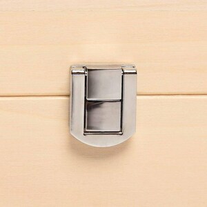 May include: Close-up of a silver-colored metal latch on a light-colored wooden surface. The latch is rectangular with a curved bottom and is centered on the wooden panel. The wood grain is visible.
