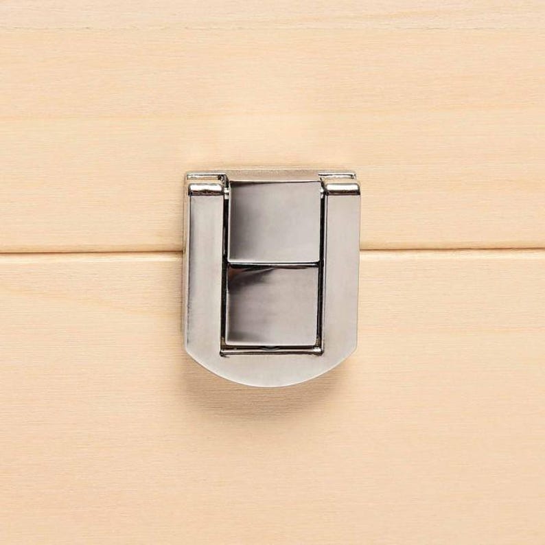 May include: Close-up of a silver-colored metal latch on a light-colored wooden surface. The latch is rectangular with a curved bottom and is centered on the wooden panel. The wood grain is visible.