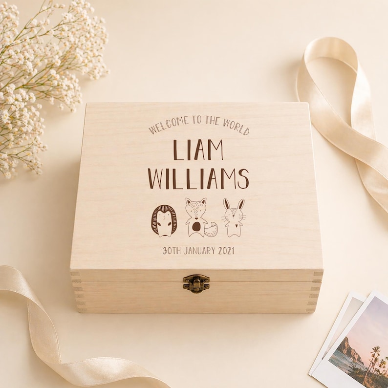May include: A light-colored wooden keepsake box with the text "WELCOME TO THE WORLD" and the name "LIAM WILLIAMS" in brown lettering. The box features illustrations of a superhero, a fox, and a rabbit. The date "30TH JANUARY 2021" is also printed on the box.