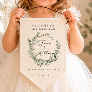 May include: A rectangular cream-colored fabric banner with a pointed bottom edge, featuring the text "WELCOME TO OUR WEDDING", "Jane & Anthony", "ST MARY'S CHURCH, BATH", and "12.04.25" printed in black, surrounded by a green eucalyptus leaf wreath.