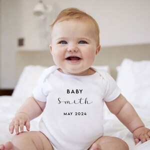 May include: A white baby onesie with black text that reads "BABY Smith MAY 2024". A baby is wearing the onesie and smiling.
