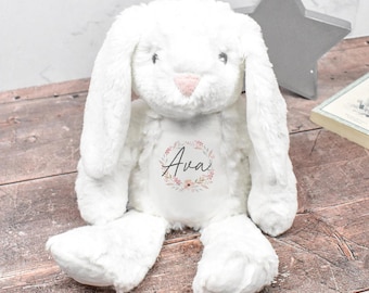 May include: A plush white stuffed bunny with long floppy ears and a soft pink nose. The center of the bunny's chest features a printed floral wreath design surrounding the name Ava in a dark script font, creating a soft and textured appearance.