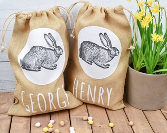 May include: Two brown burlap gift bags with white bunny illustrations and the names "George" and "Henry" printed on them. The bags are sitting on a wooden surface with yellow and white candy eggs scattered around them.