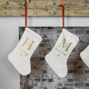May include: Two white Christmas stockings with gold lettering hanging from a wooden mantelpiece. The stocking on the left has the letter "H" and the name "harry" printed on it. The stocking on the right has the letter "M" and the name "millie" printed on it.