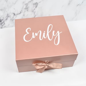 May include: A rose gold gift box with a satin ribbon and the name "Emily" printed in white on the lid.