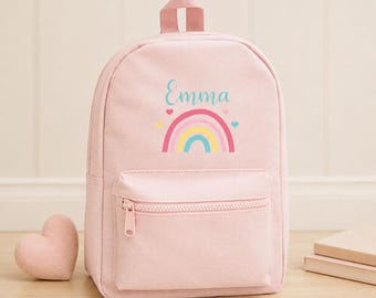 May include: A light pink backpack featuring a top handle, a front zippered pocket, and a central graphic of a rainbow with the name Emma printed in teal script above it, accented by small pink and blue hearts on a solid light pink background.