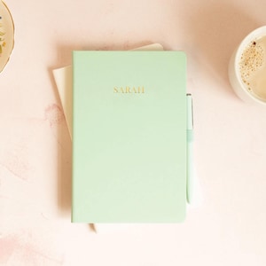 May include: A pale green notebook with the name "SARAH" in gold lettering. A matching pen rests beside the notebook, which is on top of a white sheet of paper. A cup of coffee is in the background.