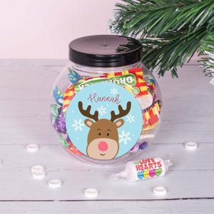 May include: A clear plastic jar filled with colorful candies, decorated with a blue and white sticker featuring a cartoon reindeer and the name "Hannah".