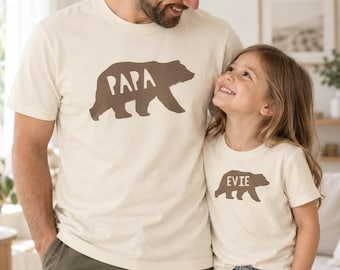 May include: Two cream-colored short-sleeve t-shirts. The larger shirt features a brown bear silhouette with the text "PAPA" inside. The smaller shirt features a brown bear silhouette with the text "EVIE" inside. Both shirts have a crew neckline.