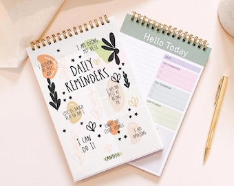 May include: Two spiral-bound notebooks with gold wire binding. The top notebook features the text Daily Reminders and various affirmations like I am doing my best. The second notebook is a planner with the text Hello Today and sections for priorities.