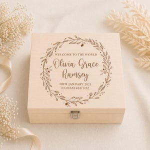 May include: A light-coloured wooden keepsake box with a decorative wreath and the text "WELCOME TO THE WORLD" and a name, date, time, and weight. The box has a metal clasp.