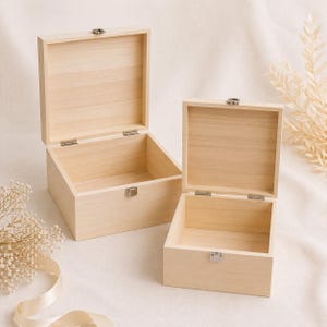 May include: Two open, unfinished wooden boxes of different sizes. The boxes have hinged lids and metal clasps. The wood grain is visible, and the boxes sit on a light-colored surface. Decorative elements are in the background.