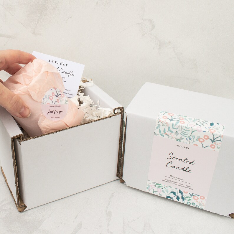 May include: Two white cardboard boxes with floral patterned labels. The box on the left is open and contains a pink scented candle wrapped in tissue paper. The label on the box on the right reads "Amy Lucy Scented Candle Hand Poured Soy (120g) Pure Soy Wax Vegan Natural Coconut & Kapok Fibre"