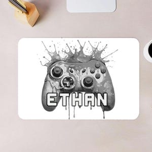 May include: A white rectangular gaming mousepad featuring a grayscale illustration of a video game controller with paint splatter effects. The name "ETHAN" is displayed in pixelated text below the controller.