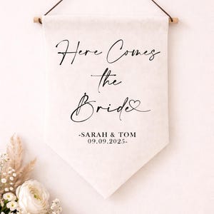 May include: A cream-colored wedding banner with the text "Here Comes the Bride" in elegant script. Below, it reads "-SARAH & TOM 09.09.2025-". The banner is suspended from a wooden dowel and a brown string. Floral arrangement in the lower left.