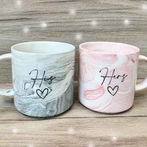 Puede incluir: Dos tazas de cer&aacute;mica con patrones marmoleados. Una taza es gris con la palabra "His" y un coraz&oacute;n. La otra taza es rosa con la palabra "Hers" y un coraz&oacute;n.