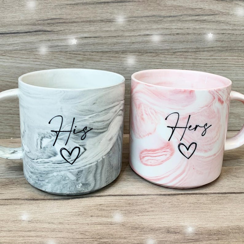 Puede incluir: Dos tazas de cer&aacute;mica con patrones marmoleados. Una taza es gris con la palabra "His" y un coraz&oacute;n. La otra taza es rosa con la palabra "Hers" y un coraz&oacute;n.