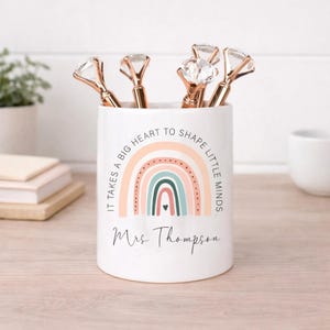 May include: White ceramic pen holder with a rainbow design and the text "It takes a big heart to shape little minds" and "Mrs Thompson". The holder contains several pens with diamond-shaped tops.