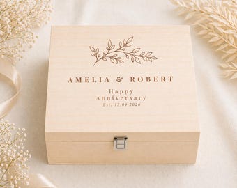 May include: A light-colored wooden box with a hinged lid and a silver-colored clasp. The box is engraved with the names "AMELIA & ROBERT", a floral design, and the words "Happy Anniversary Est. 12.09.2026".