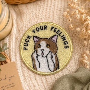 Può includere: Una toppa ricamata circolare con sfondo giallo chiaro raffigurante un gatto marrone e bianco con occhi spalancati e bocca aperta. Il gatto solleva entrambe le zampe per mostrare i medi. Il testo FUCK YOUR FEELINGS è ricamato in nero.