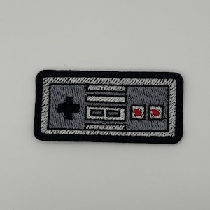 May include: A rectangular embroidered patch featuring a gray video game controller design with a black border. The design includes a black directional pad on the left, two small red buttons on the right, and white detailing representing the console buttons.