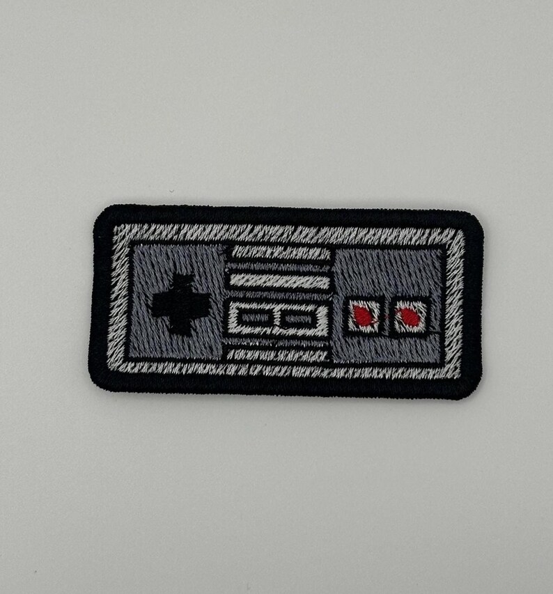 May include: A rectangular embroidered patch featuring a gray video game controller design with a black border. The design includes a black directional pad on the left, two small red buttons on the right, and white detailing representing the console buttons.