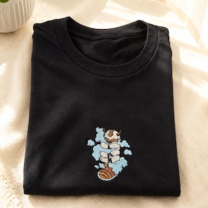 Può includere: Una t-shirt nera a girocollo con un piccolo ricamo centrato raffigurante una creatura bianca e pelosa con corna e segni marroni, circondata da nuvole azzurre e una coda marrone, posizionata sulla parte inferiore del petto.