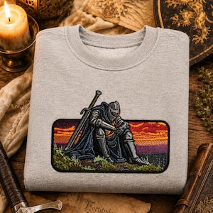 Puede incluir: Una sudadera gris claro de cuello redondo con un parche rectangular bordado en el pecho. El parche muestra a un caballero con armadura plateada y capa azul oscuro sentado en una colina con una espada, frente a un cielo de atardecer.