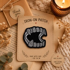 May include: An iron-on patch featuring a black and white embroidered design of sharp vampire fangs. The patch is mounted on a rectangular tan card with the text IRON-ON PATCH, MADE WITH CARE - FOR YOU, and EASY TO APPLY LONG LASTING QUALITY.
