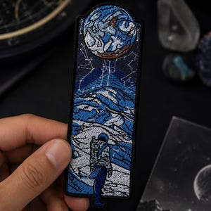 May include: Embroidered patch featuring an astronaut gazing at a celestial landscape. The design includes a blue and white planet, a starry sky, and a detailed depiction of a mountainous terrain. The patch is held by a hand.