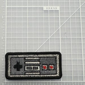 May include: A rectangular embroidered patch featuring a gray video game controller design with a black border, a black directional pad, two white buttons, and two red circular buttons, positioned on a grid mat marked 2.5 x 3.5 inches.