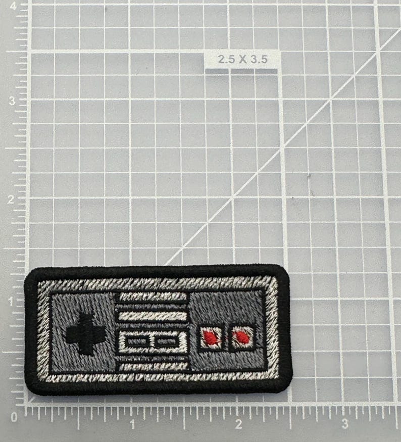 May include: A rectangular embroidered patch featuring a gray video game controller design with a black border, a black directional pad, two white buttons, and two red circular buttons, positioned on a grid mat marked 2.5 x 3.5 inches.